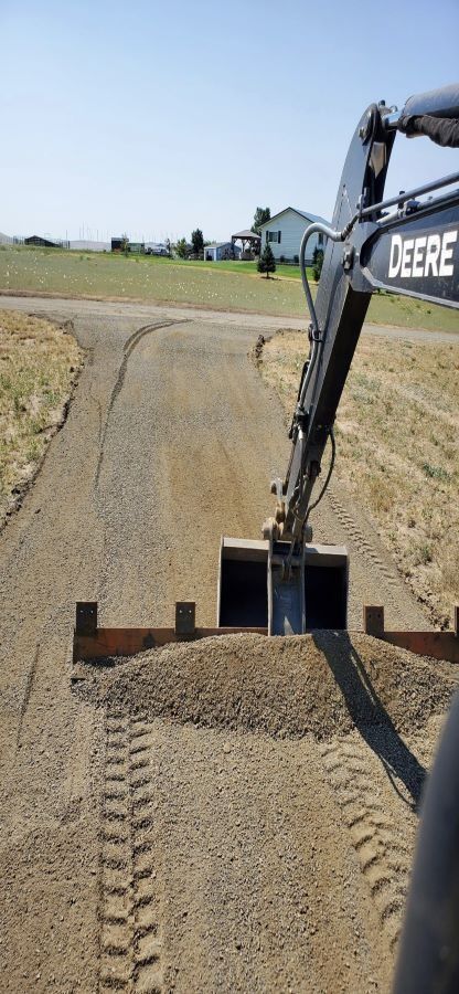 A Deere excavator grading a gravel driveway on a sunny day, with a house in the background.