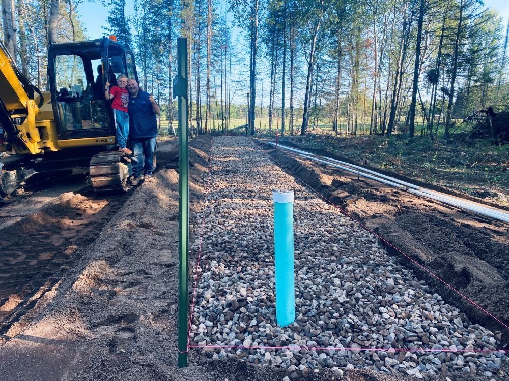 Construction site with backhoe, two people, gravel trench, and blue pipe. Forest backdrop.