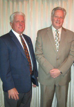 Two men in formal suits stand side-by-side against a draped curtain background.