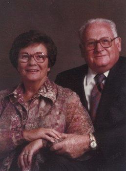 A portrait of two people posing together, both wearing glasses, with hands gently resting on each other in front.