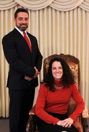 A smiling person in a suit and red tie stands behind a person in a red turtleneck sitting in an ornate chair.