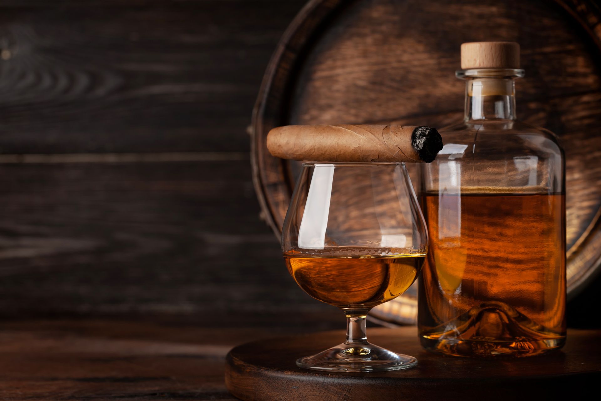 Bicchiere di liquido ambrato, bottiglia e sigaro acceso su un vassoio di legno, sullo sfondo una botte scura.