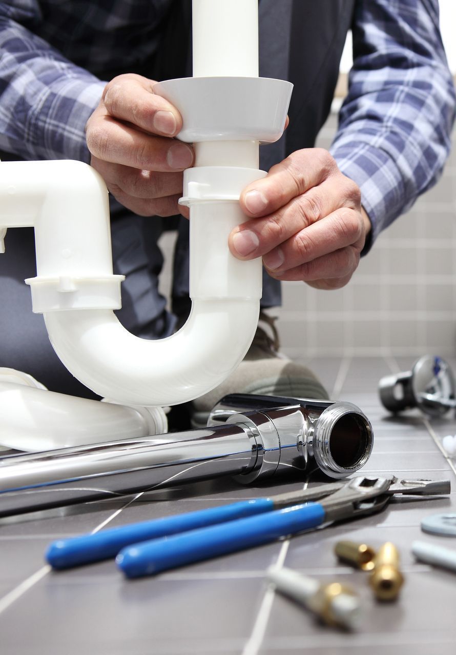 Plombier connectant un siphon en P en plastique blanc sous un évier avec des outils à proximité, sol carrelé.