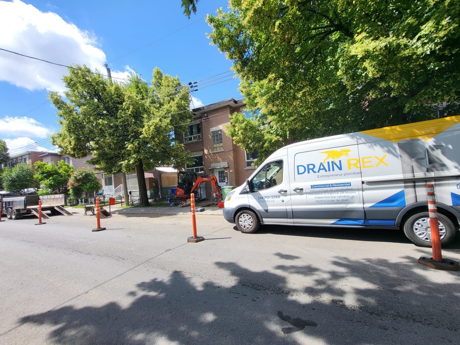 Une camionnette Drain Flex est garée dans une rue en travaux. Une excavatrice est stationnée près d'un bâtiment en briques et des cônes de signalisation sont placés le long de la route.