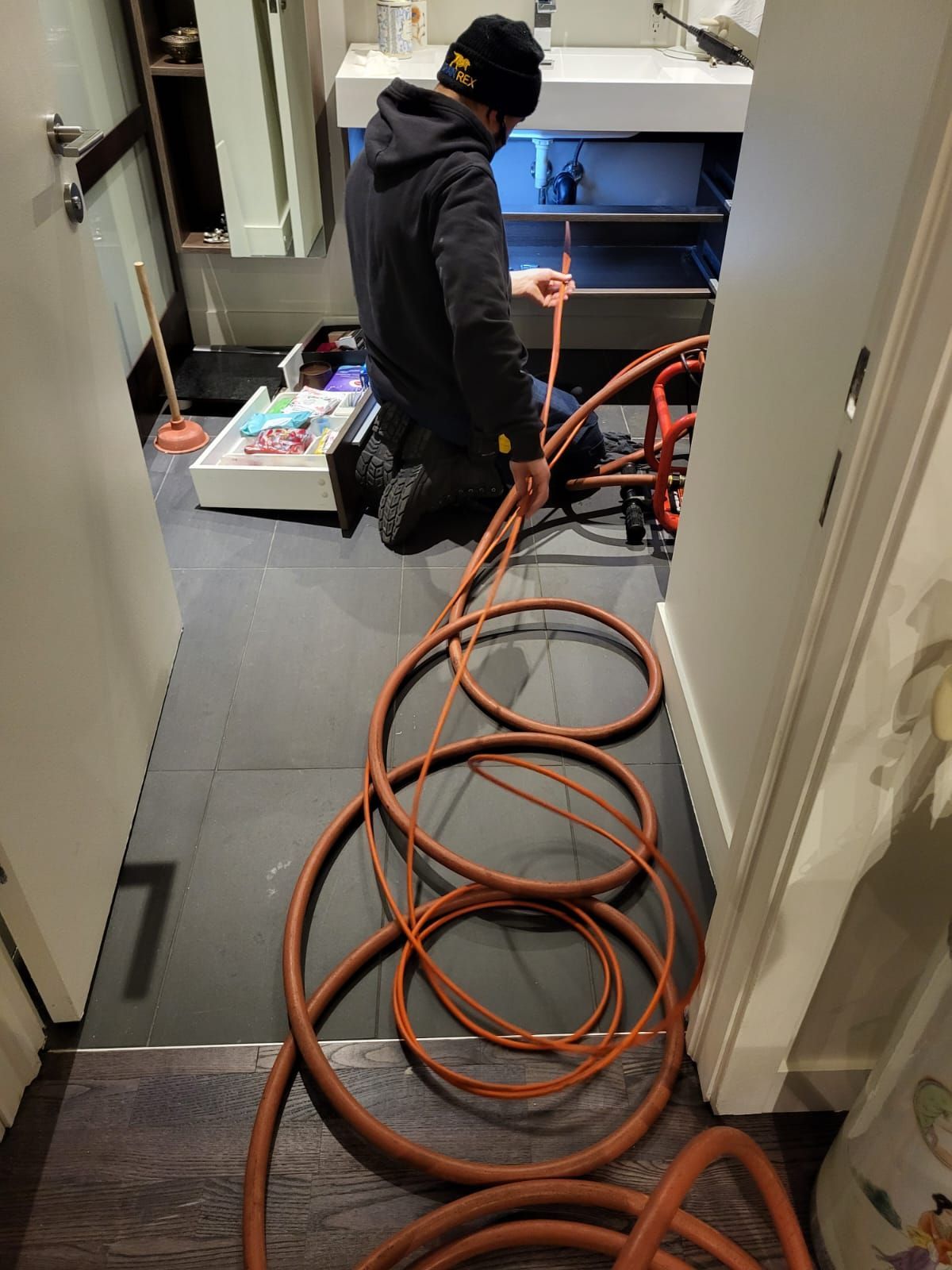 A person kneels in a bathroom working on plumbing, with coiled orange hoses in the foreground.