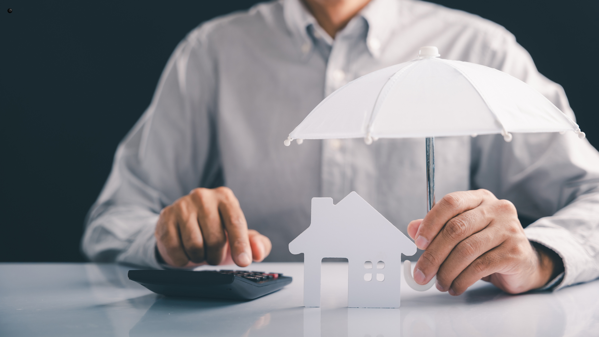 A person holding a white umbrella over a small house model, symbolizing the protection provided by homeowners insurance with a calculator in the background, suggesting financial planning.