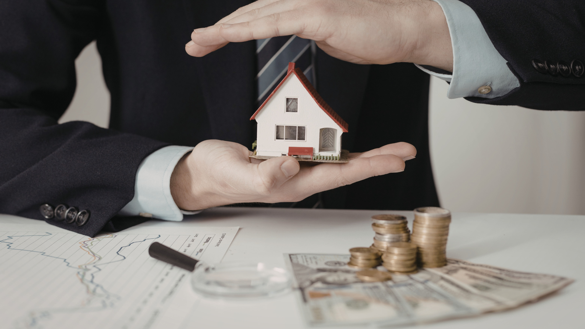 Business professional holding a small model house in one hand, surrounded by coins and dollar bills,