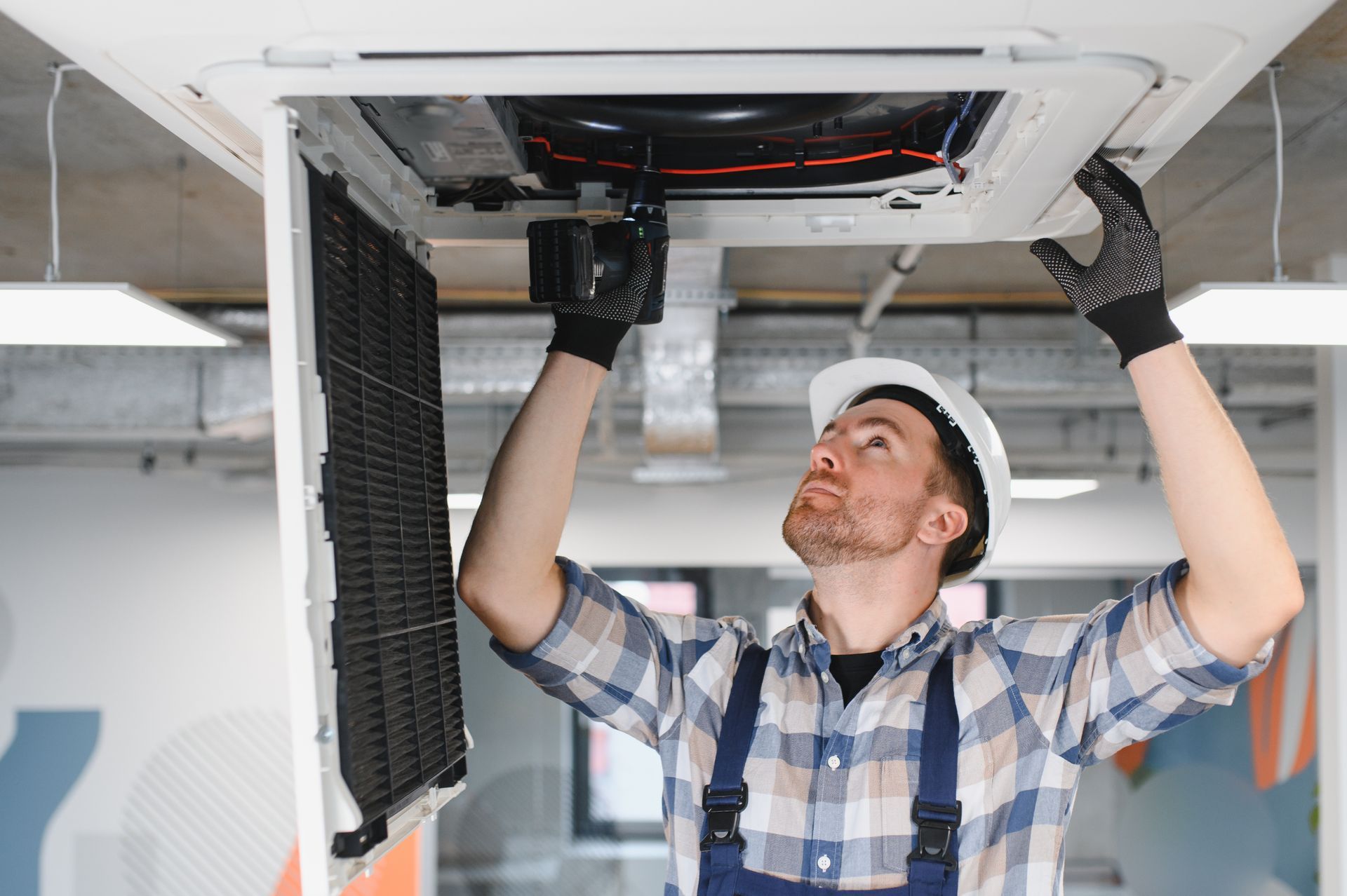 A person in a hard hat and work coveralls inspects the interior of a ceiling-mounted air conditioning unit.