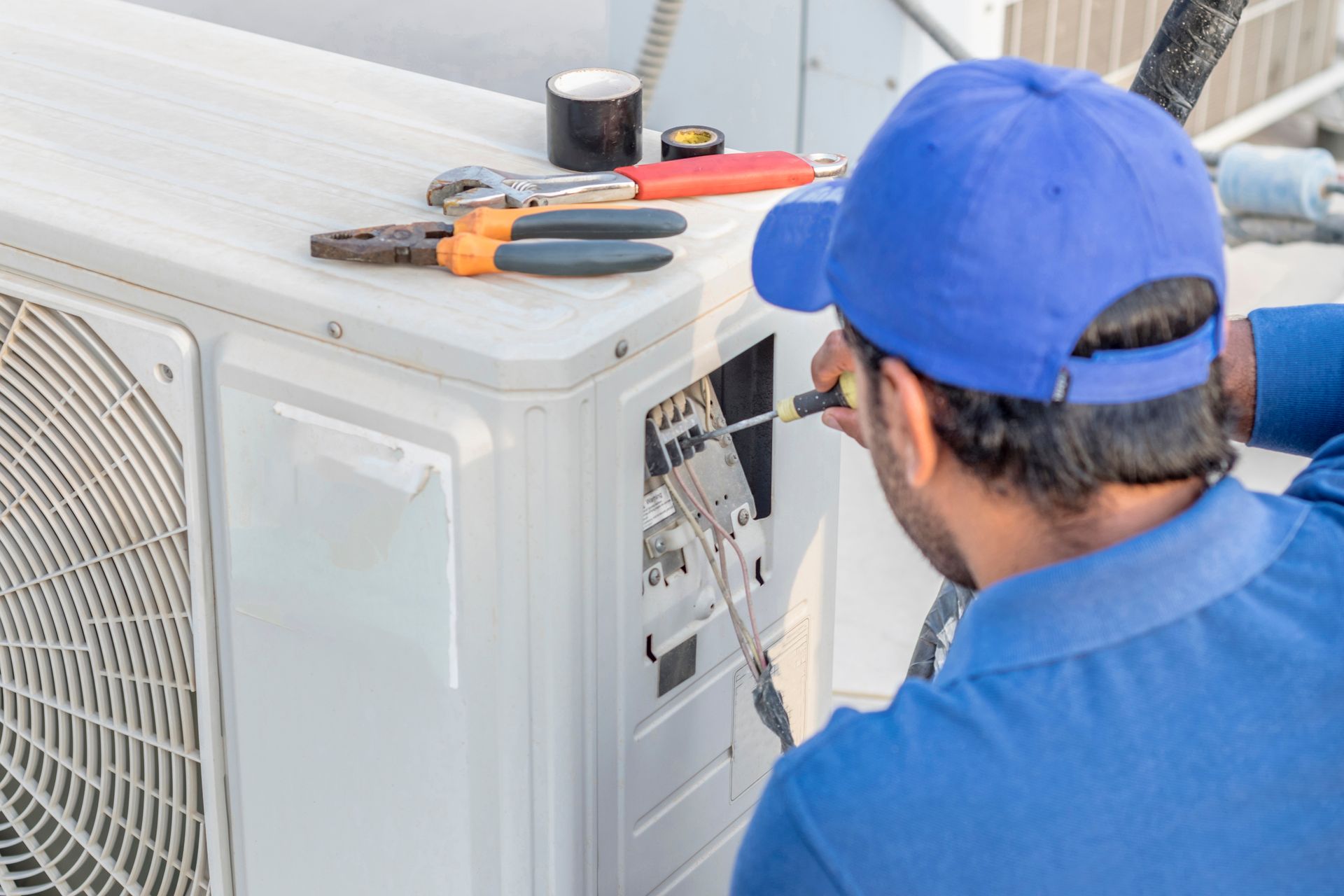 A technician wearing a blue uniform and cap repairs an outdoor air conditioning unit with tools nearby.