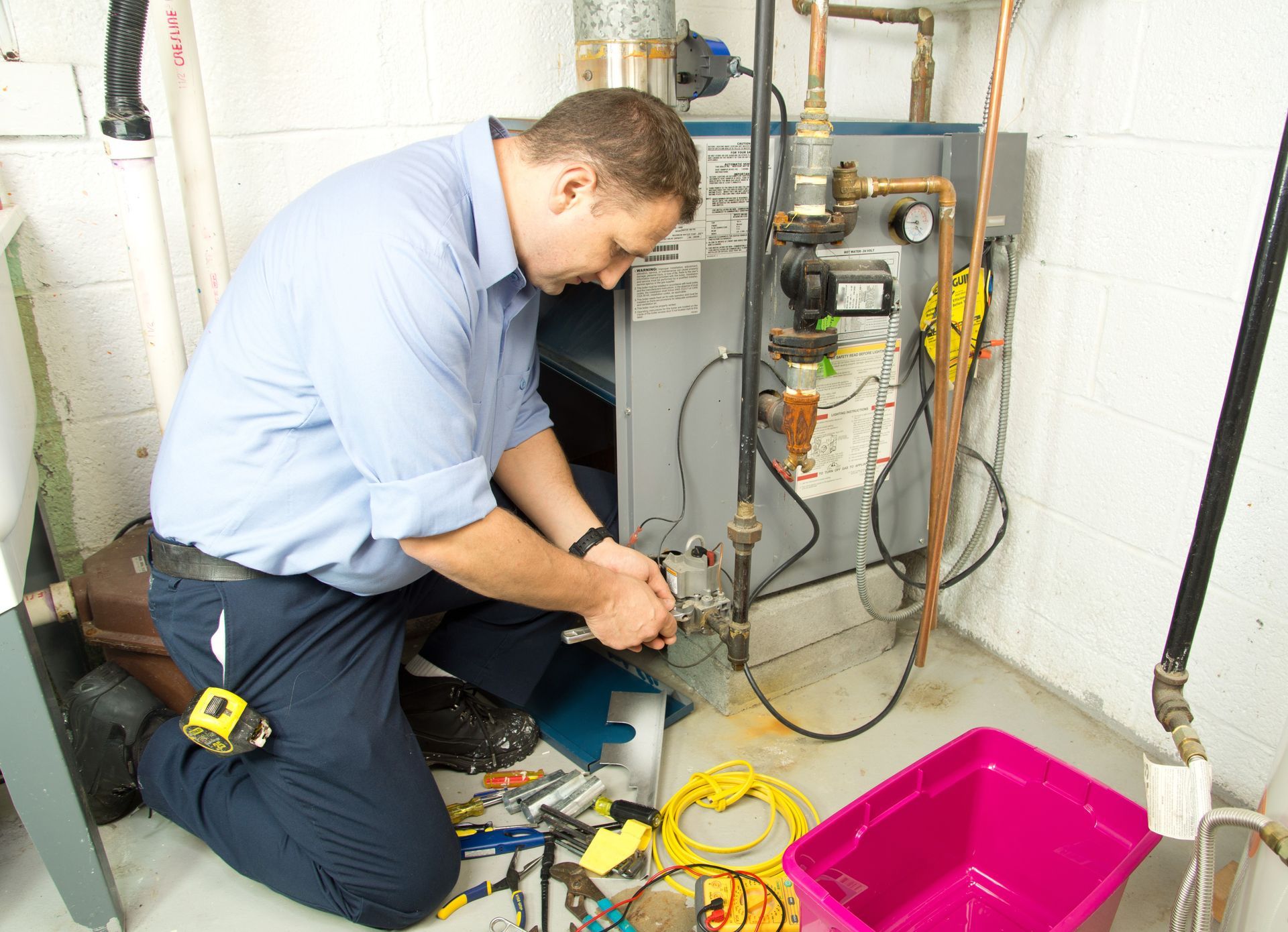 Technician kneels in a basement, repairing a residential boiler unit surrounded by tools and a pink bin.