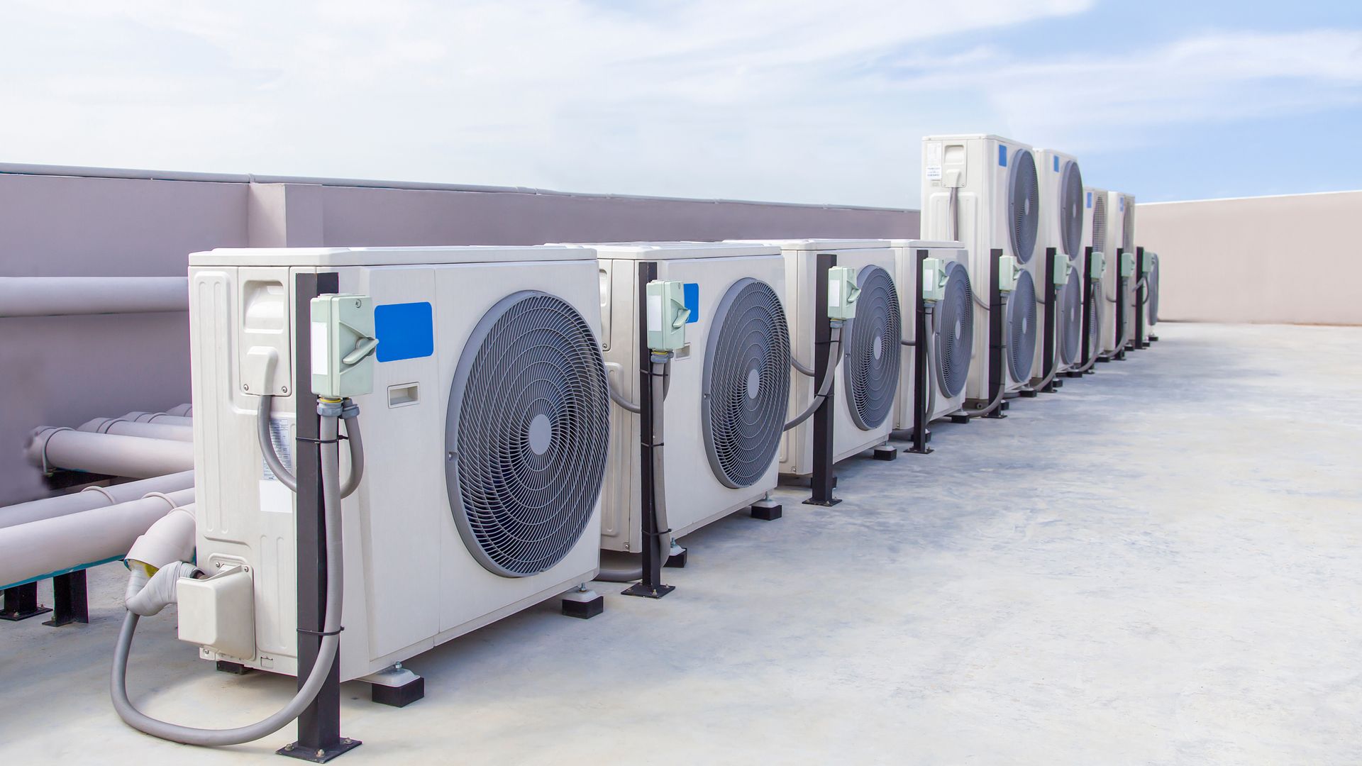 A row of beige industrial air conditioning units positioned on a flat rooftop against a clear blue sky.