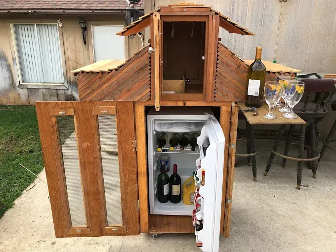 A wooden outdoor bar cabinet with a small refrigerator, shelf, and wine glasses, standing on a patio.