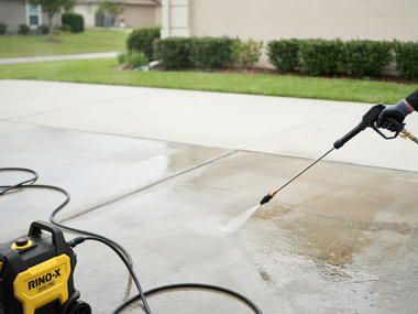Pressure washer spraying a driveway beside a house, with a yellow machine and hose in the foreground