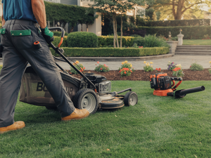 Person mowing a lawn with a push mower beside an orange leaf blower in a landscaped yard