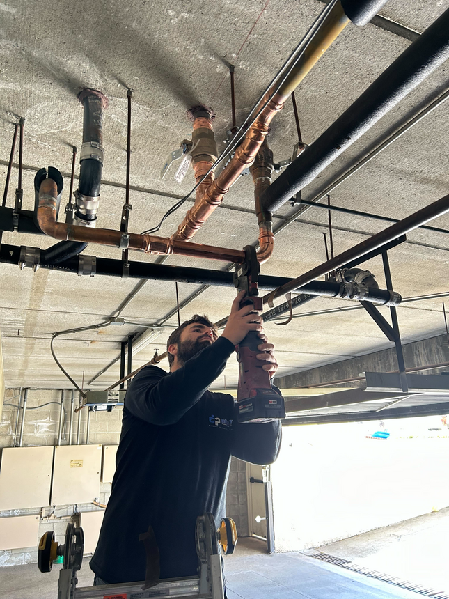 Plumber on a ladder, using a drill on copper pipes attached to a ceiling in a garage.