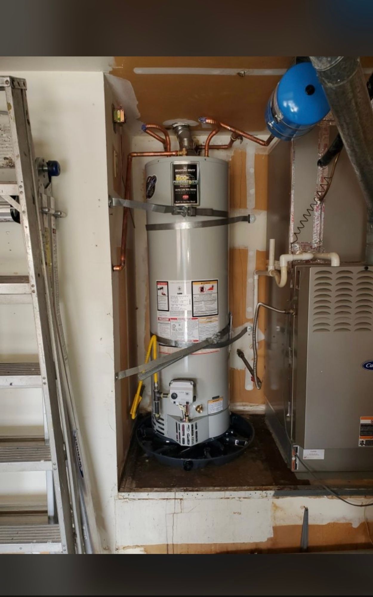 a water heater is sitting in a corner of a room next to a ladder .