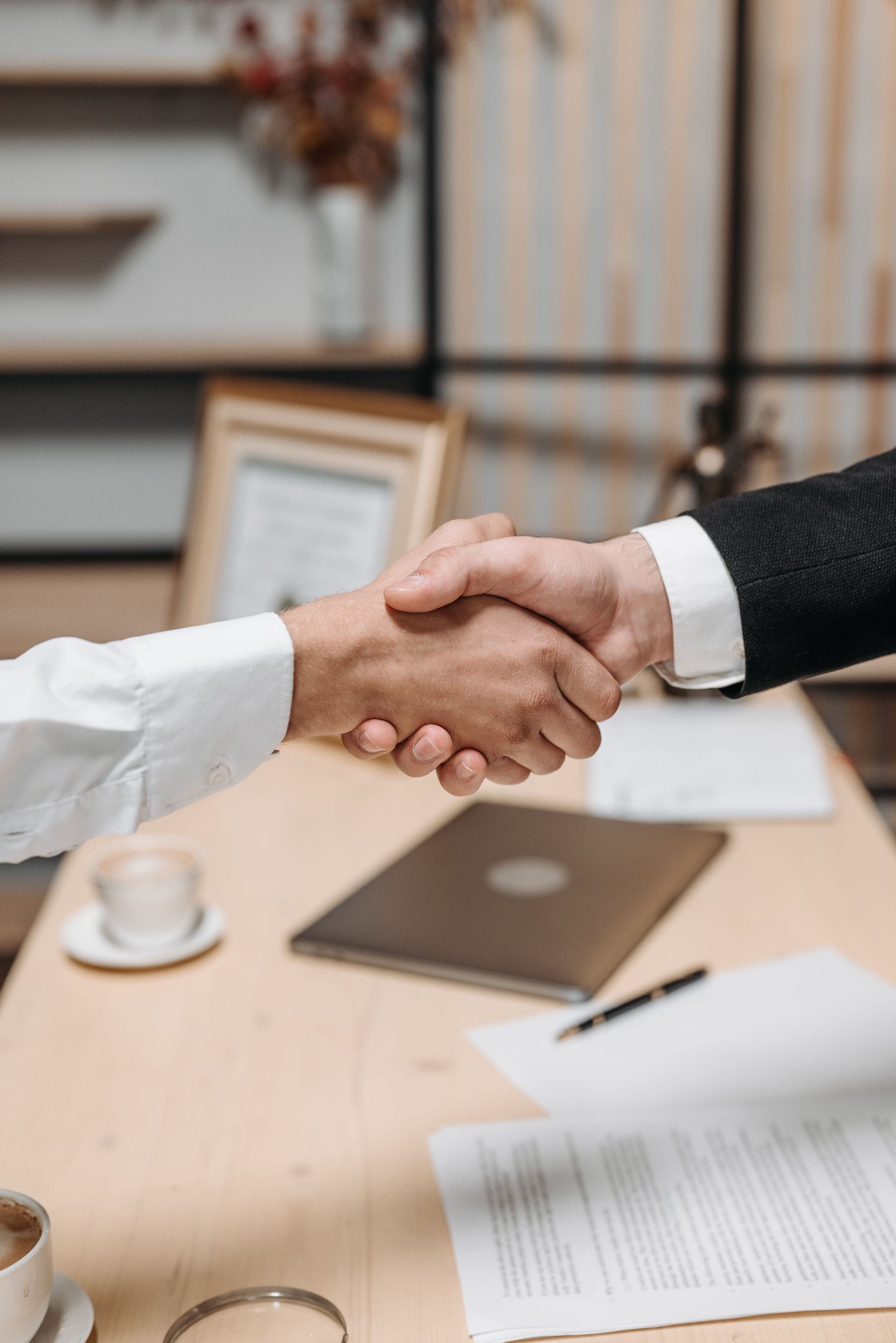 Two people shaking hands across a desk in a meeting room with documents and a laptop.