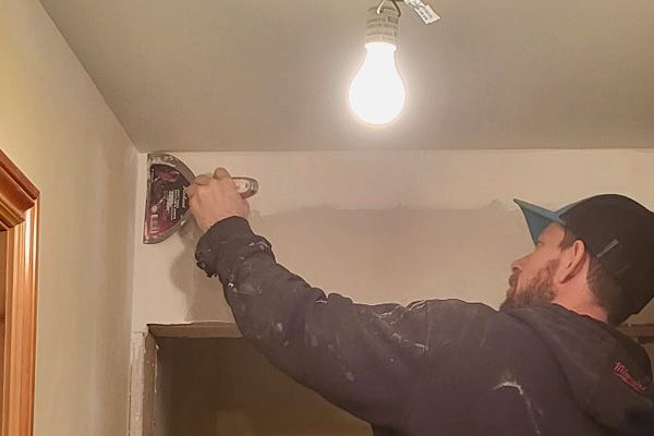 A hand uses a metal putty knife to apply white joint compound to a ceiling seam.
