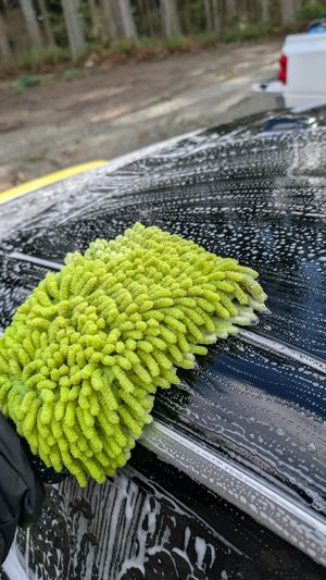 A lime-green microfiber wash mitt cleans a soapy, wet black car roof outdoors.