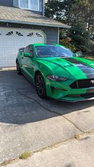 A bright green Ford Mustang with black racing stripes parked on a residential driveway in front of a house.