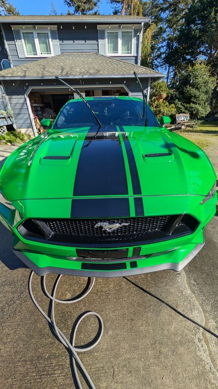 A bright green Ford Mustang with black racing stripes parked in a driveway in front of a house.