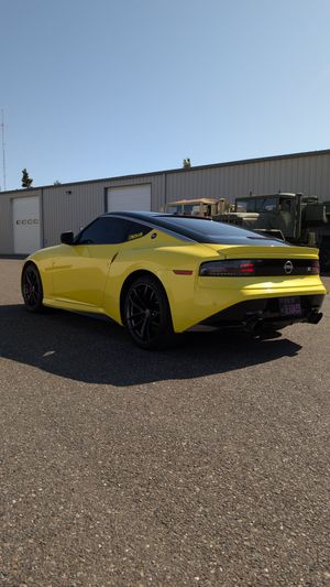 A yellow Nissan Z sports car parked on a gravel lot in front of a metal industrial building.