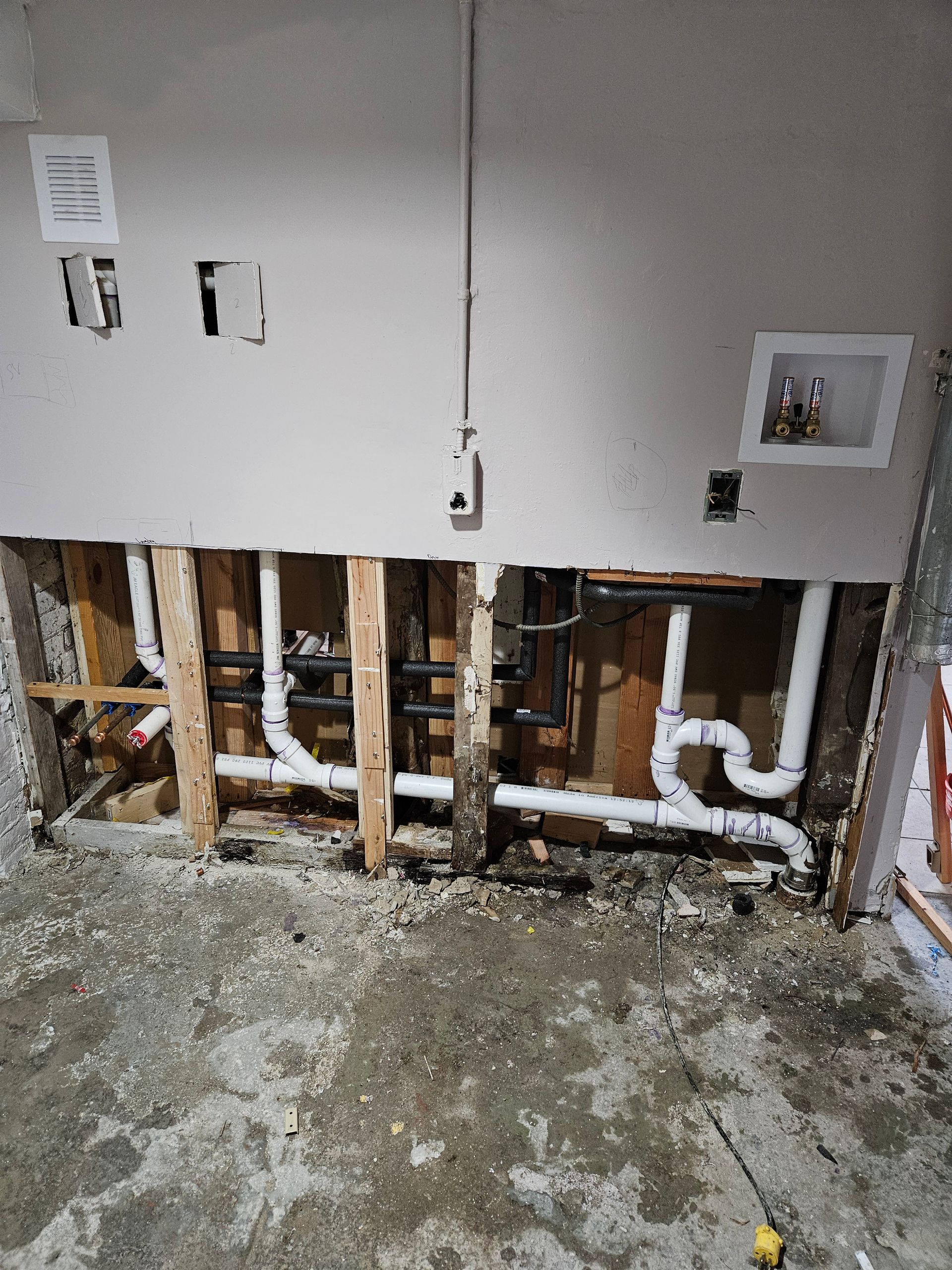 Plumbing pipes exposed in a wall during construction, with a washing machine hookup and electrical outlets.