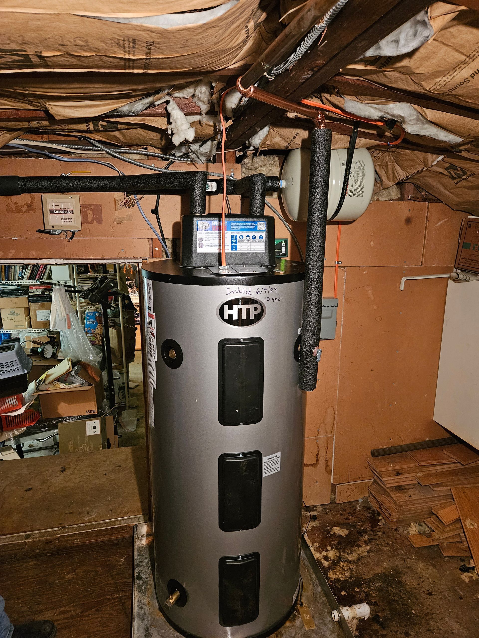 A shiny, silver HTP water heater in a dimly lit basement with exposed pipes and wood.
