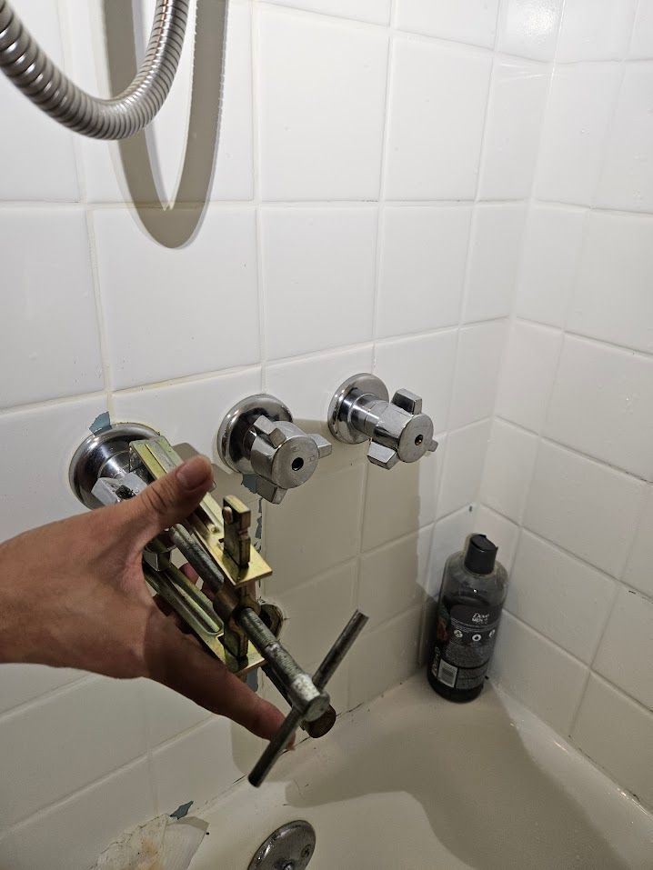A hand holding shower valve, next to a shower with two wall-mounted faucet handles.
