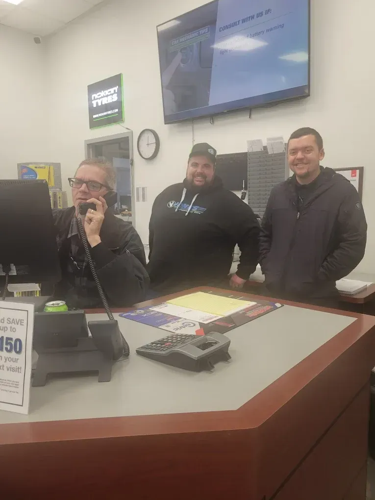 Three men stand behind a store counter, smiling in a bright electronics shop. | Jones Complete Car Care
