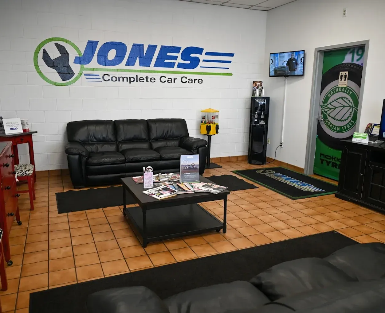 Waiting area at Jones Complete Car Care with black couches, coffee table, and TV in a tiled lobby. | Jones Complete Car Care