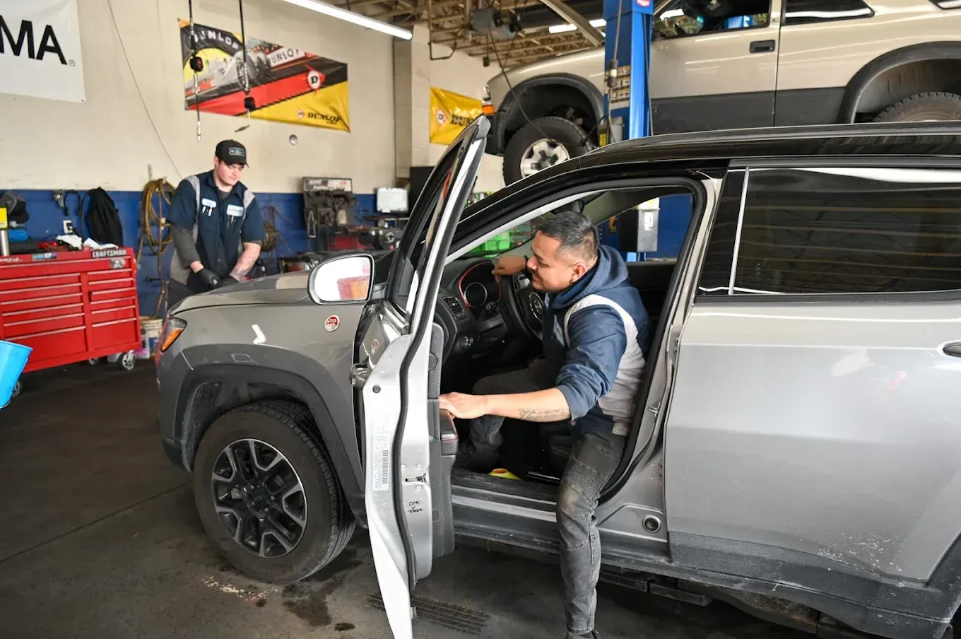 Mechanic leaning into a silver SUV in an auto shop, with a car lifted in the background. | Jones Complete Car Care