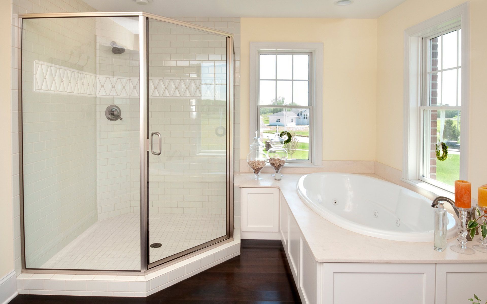 Bathroom with a glass shower, white bathtub, two windows, and dark wood floors.