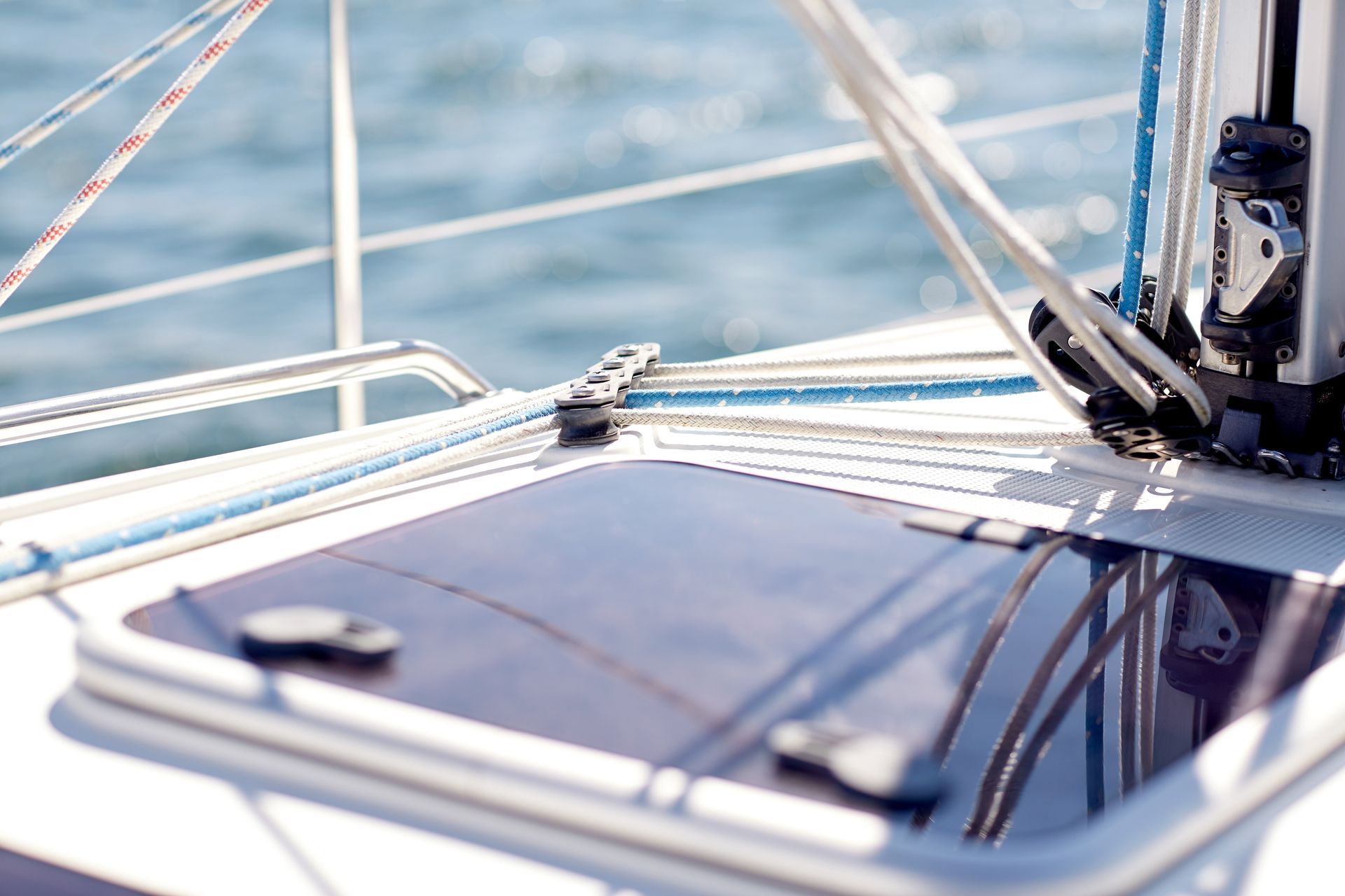 Sailboat deck detail with window, ropes, and hardware against a blurred blue sea.