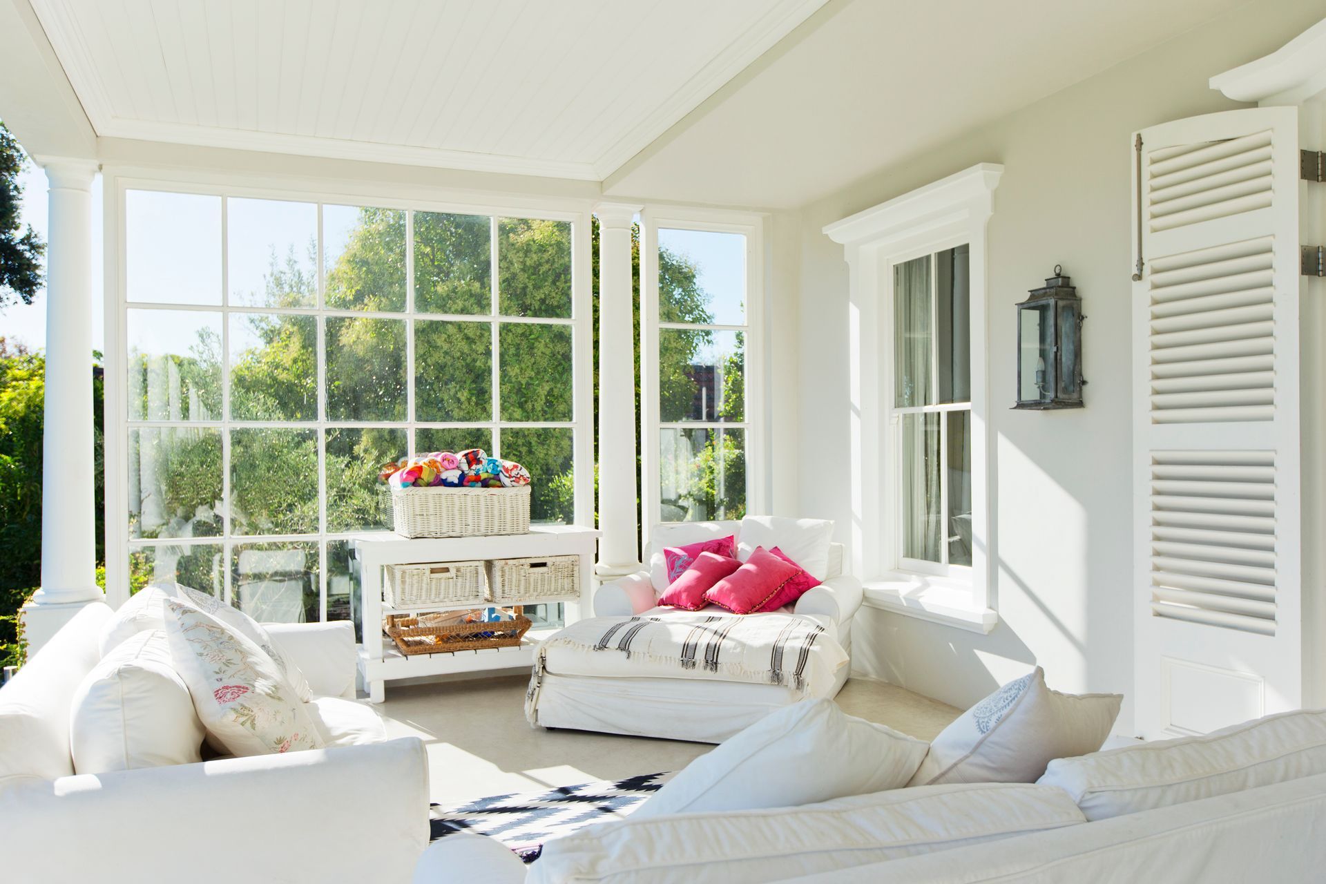 Sunroom with white furniture, large windows, and a basket of flowers overlooking greenery.