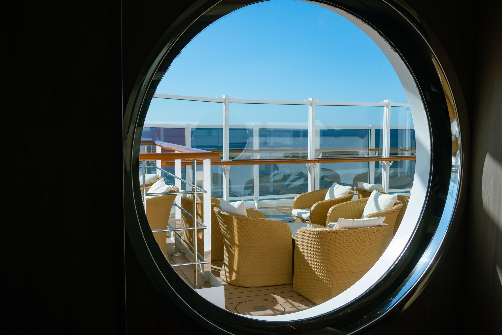Porthole view of a cruise ship deck with seating, blue sky and ocean.