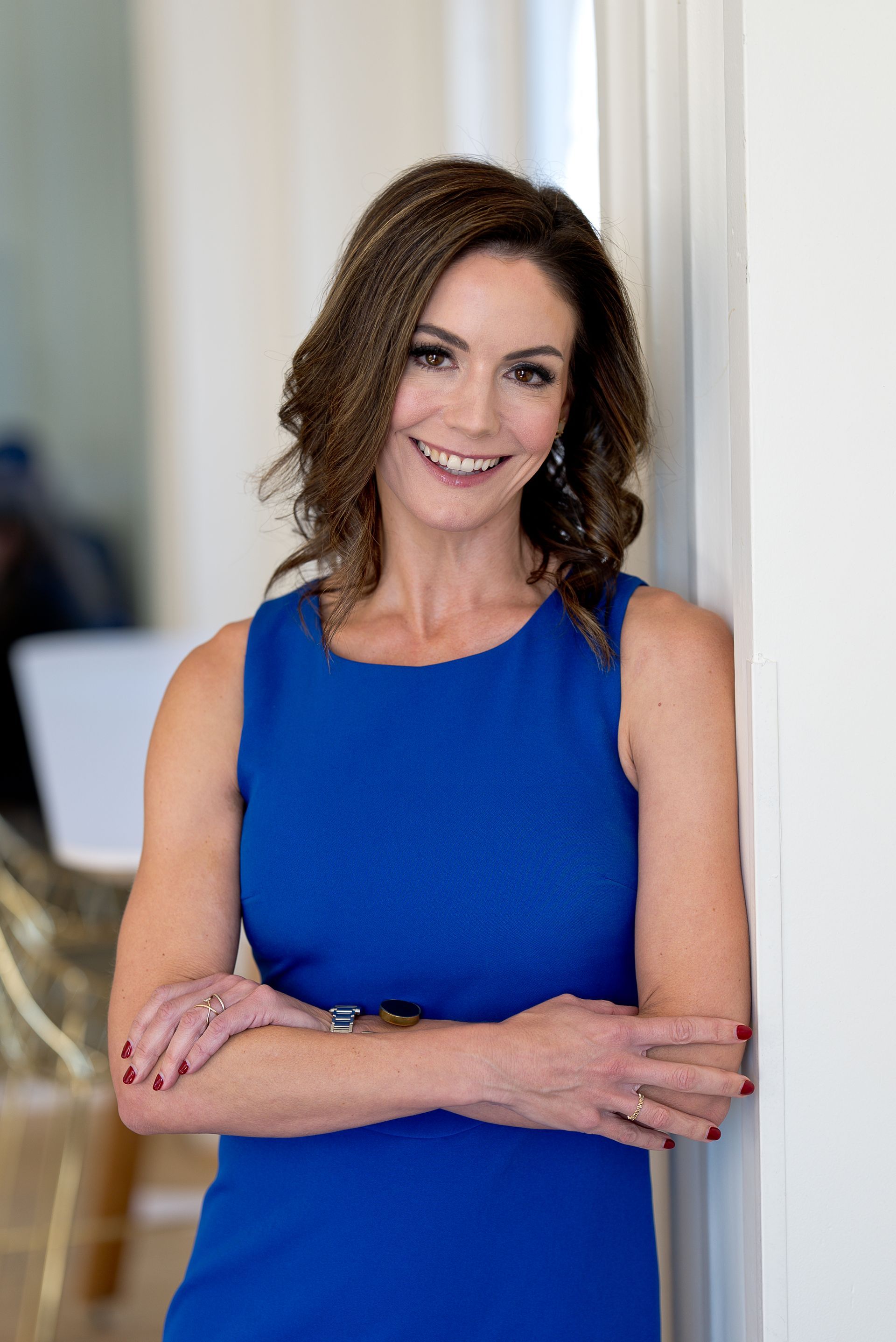 Woman with brown hair and a colorful dress smiles indoors, two mirrors behind.