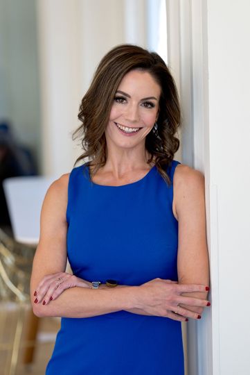 Woman with brown hair and a colorful dress smiles indoors, two mirrors behind.