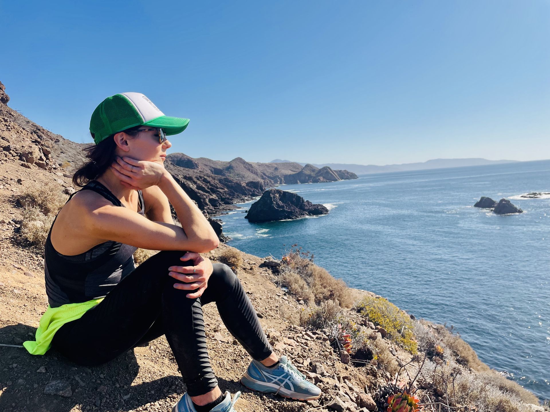 Woman in workout gear with green hat looks out at the ocean from a rocky cliff.
