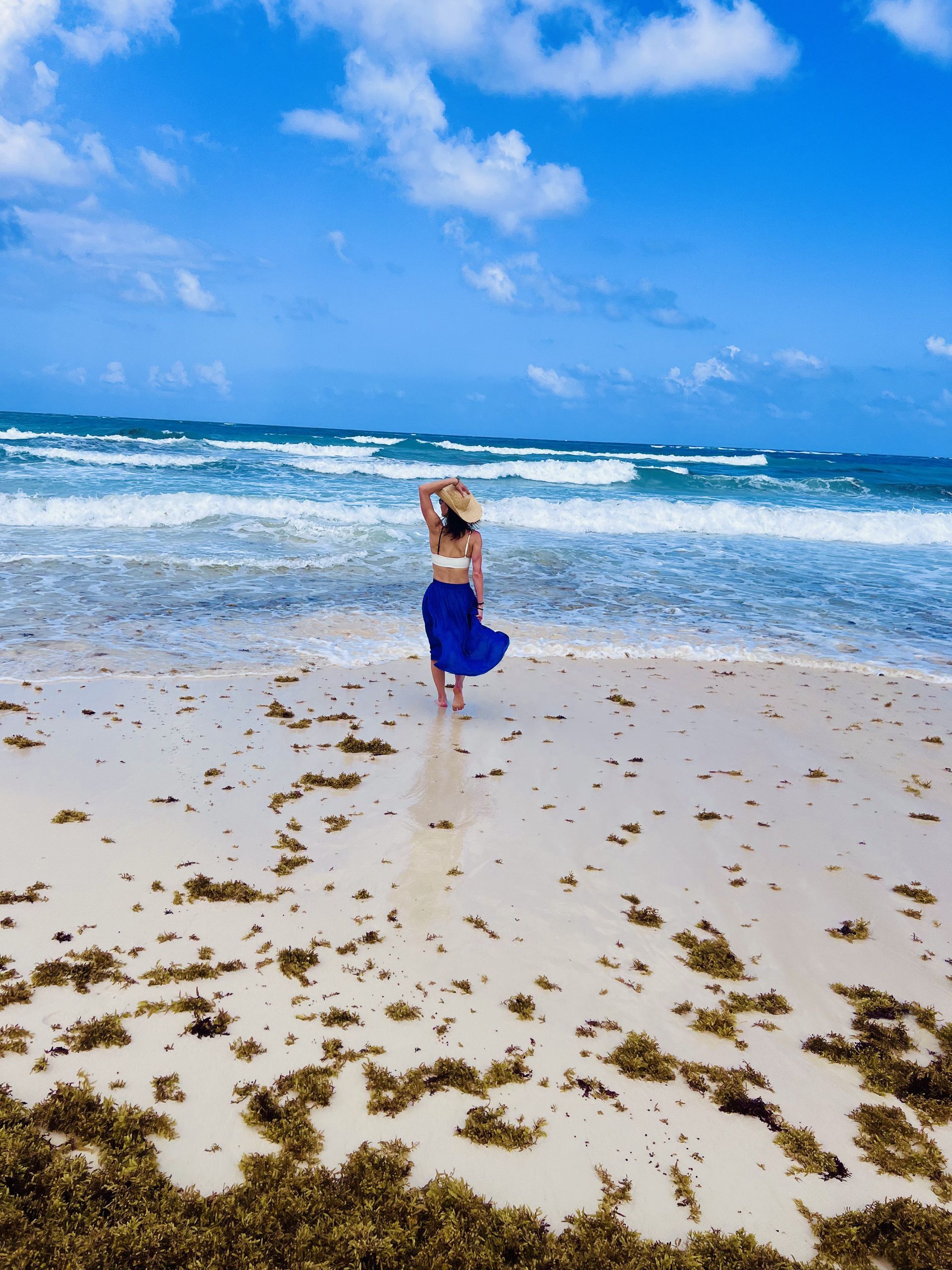 Woman in blue skirt and hat on beach, looking at ocean under a blue sky.