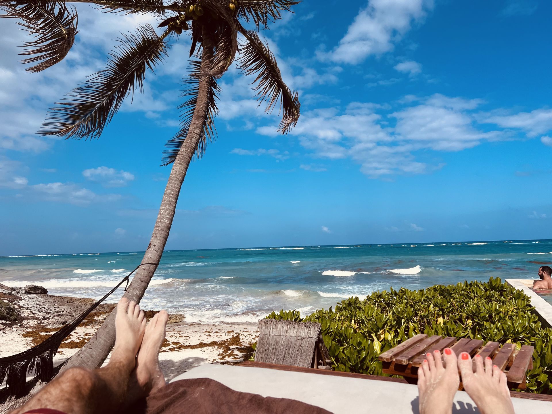 Feet relaxing on lounge chairs, beach view with palm tree, blue sky, ocean waves.