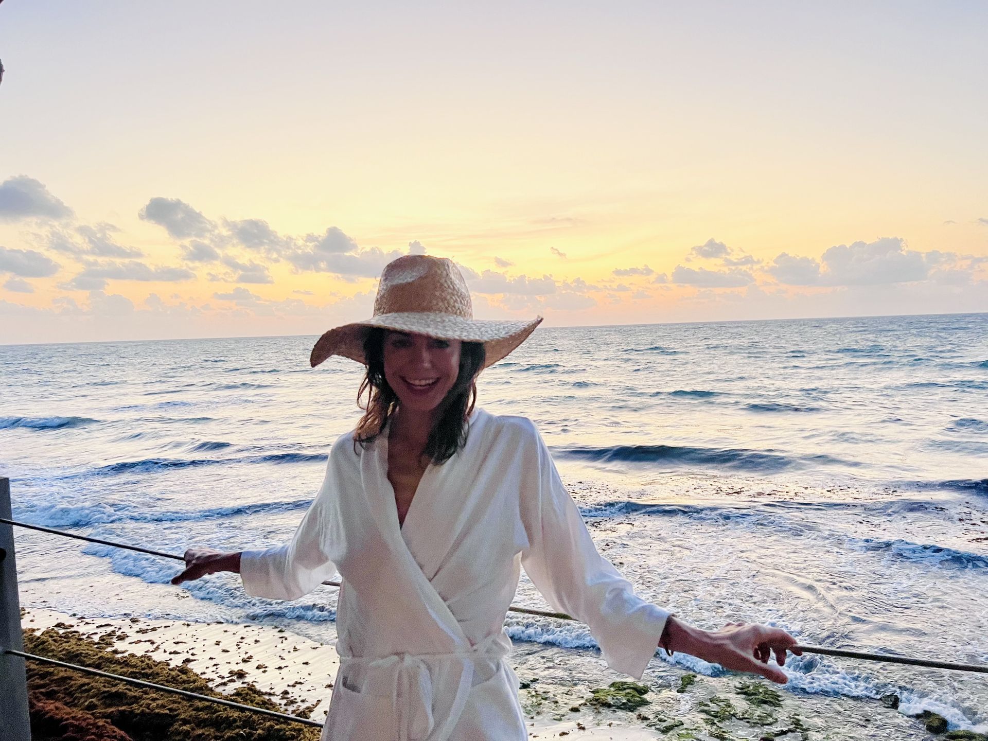 Woman in a straw hat and robe smiles at sunset over the ocean.