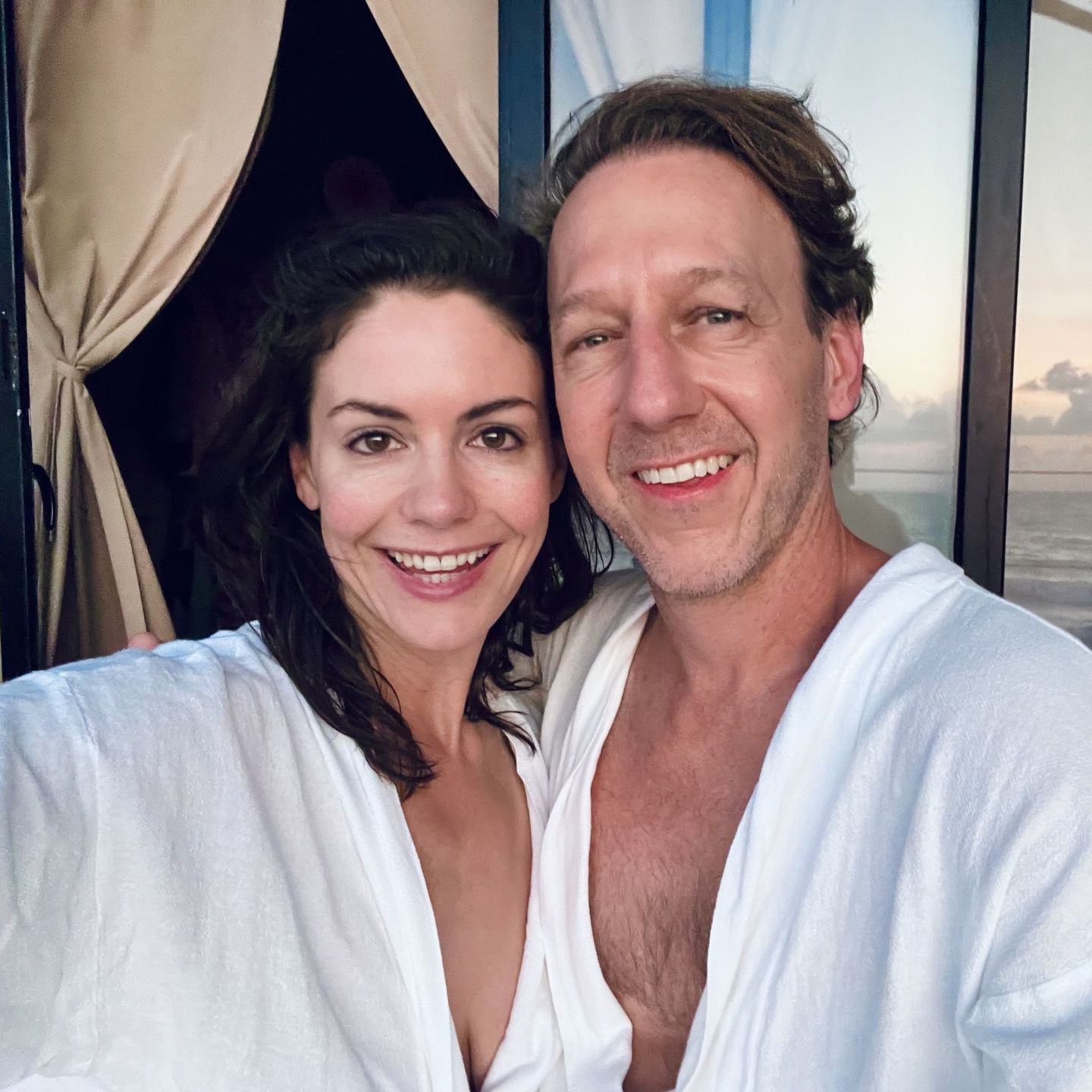 Woman and man smiling in white robes, standing on a balcony. Ocean and sunset are visible.