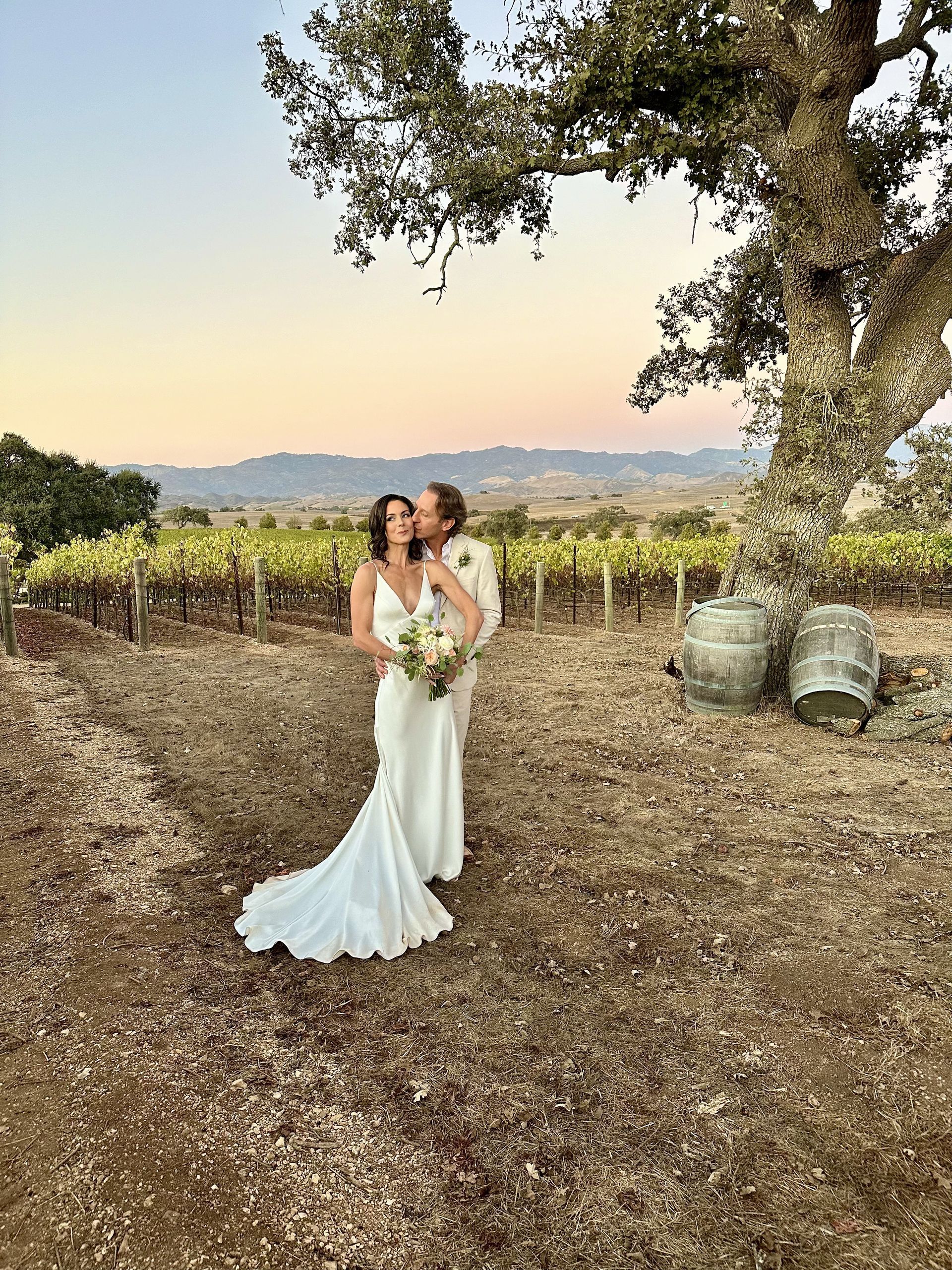 Bride and groom kissing in a vineyard at sunset. She wears a white dress, he has light hair, and they hold a bouquet.