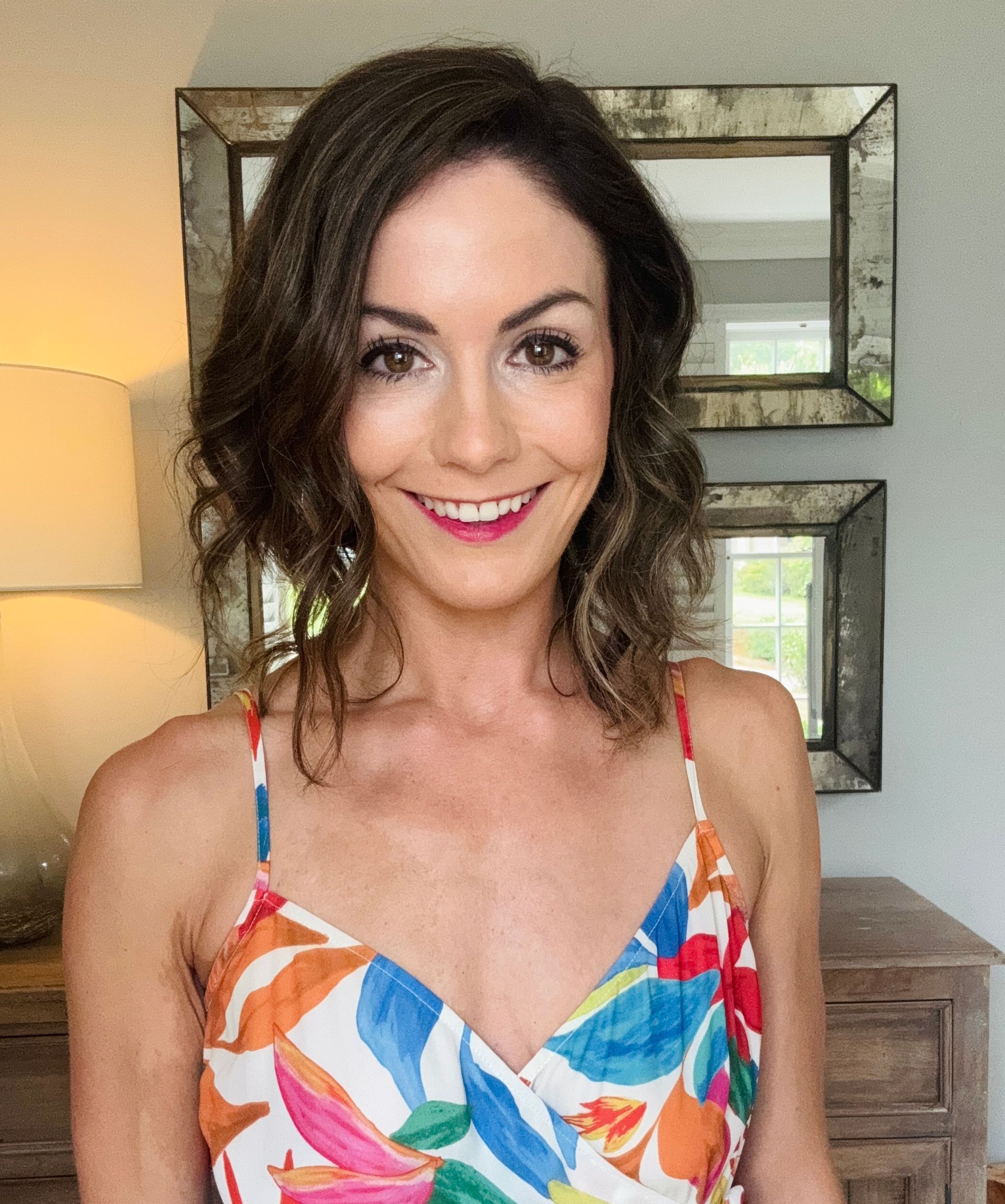 Woman with brown hair and a colorful dress smiles indoors, two mirrors behind.
