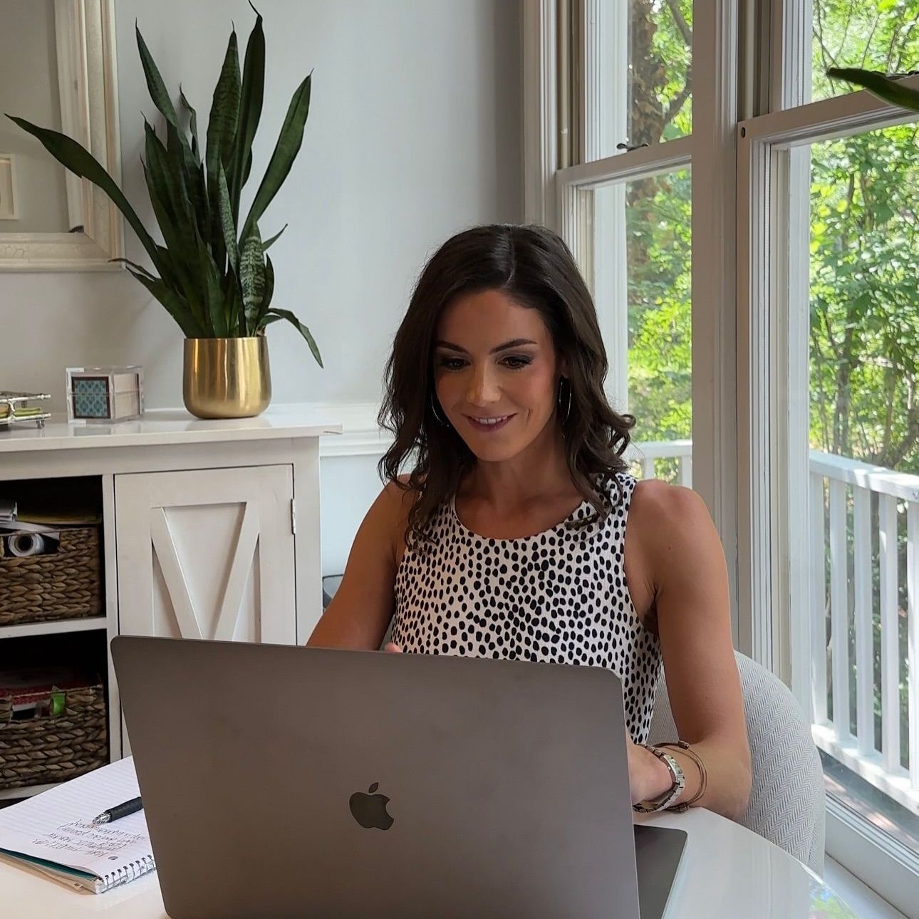 Woman working on laptop at a table by a window; plant in the background.