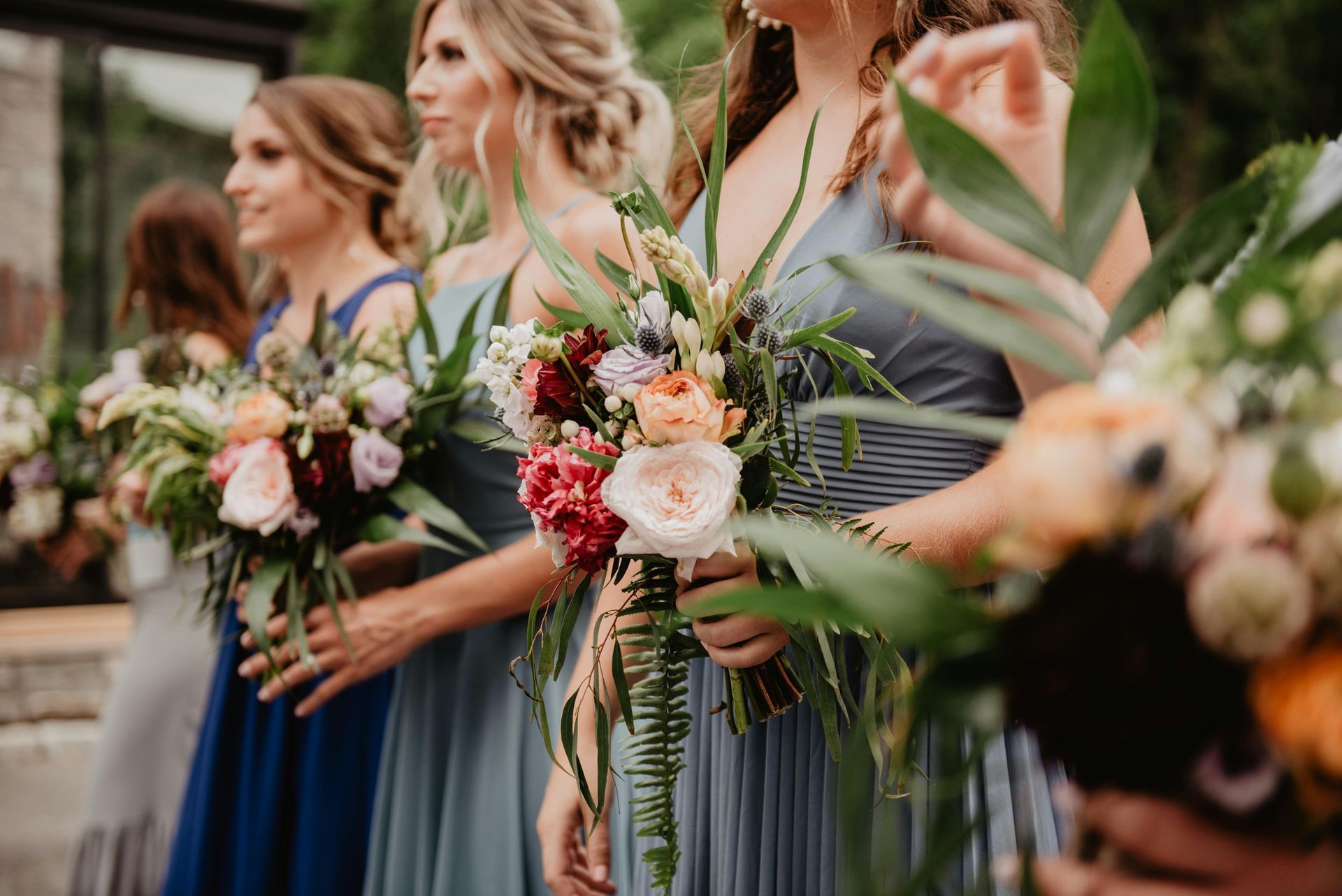 Bridesmaids in blue dresses holding colorful bouquets, standing outdoors.