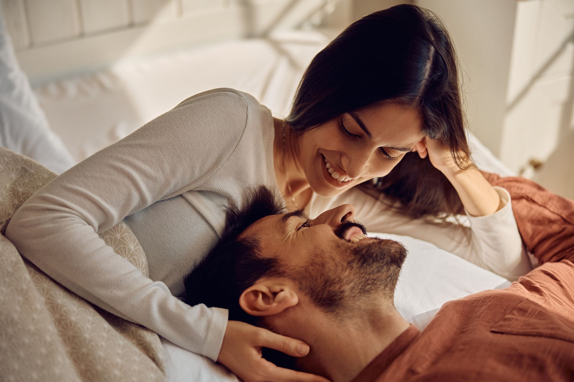 Woman smiling down at a man lying in bed. Both are in soft lighting, appearing happy.