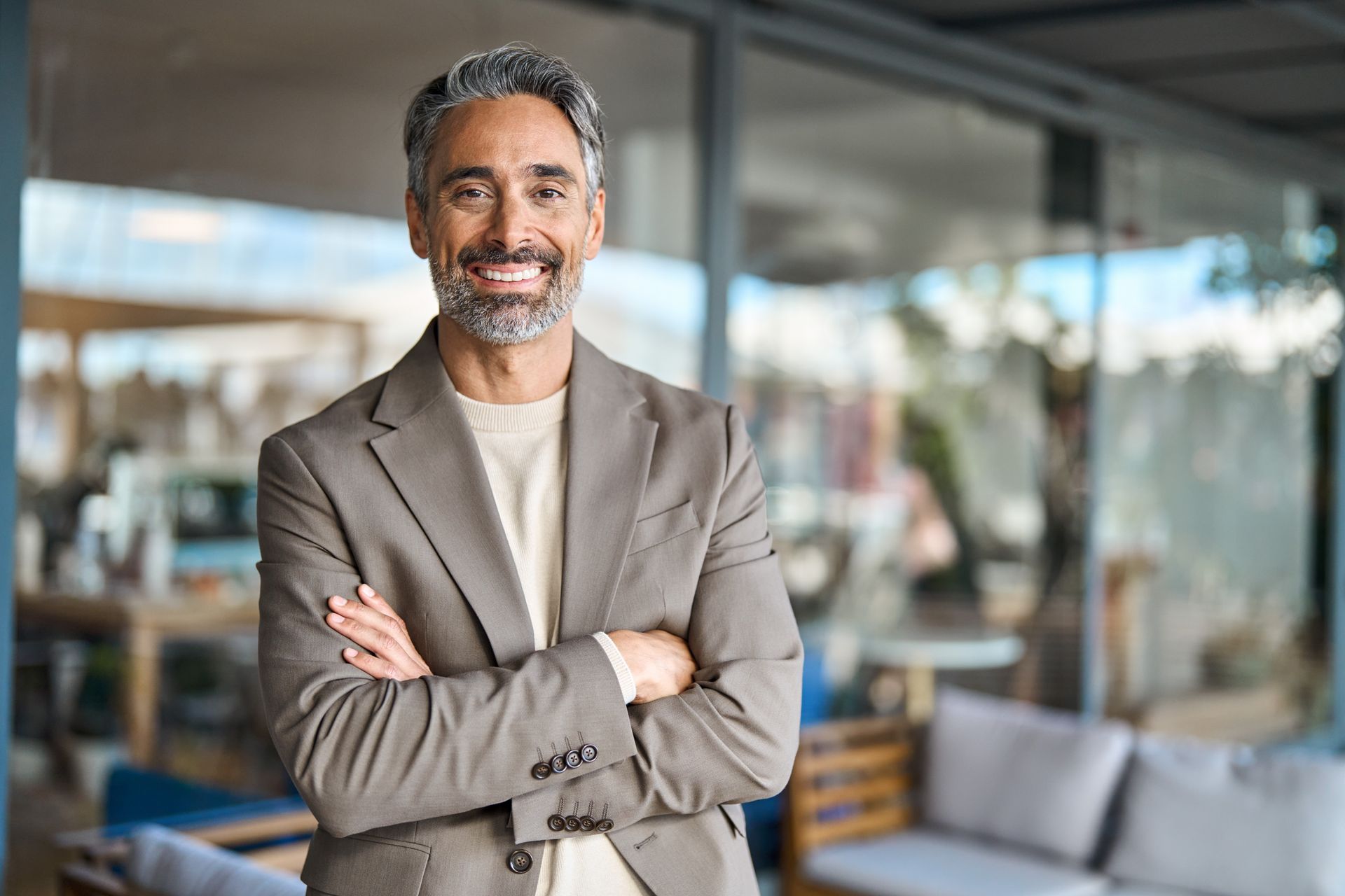 Man with graying hair and beard, smiling with arms crossed, wearing a blazer in a modern setting.