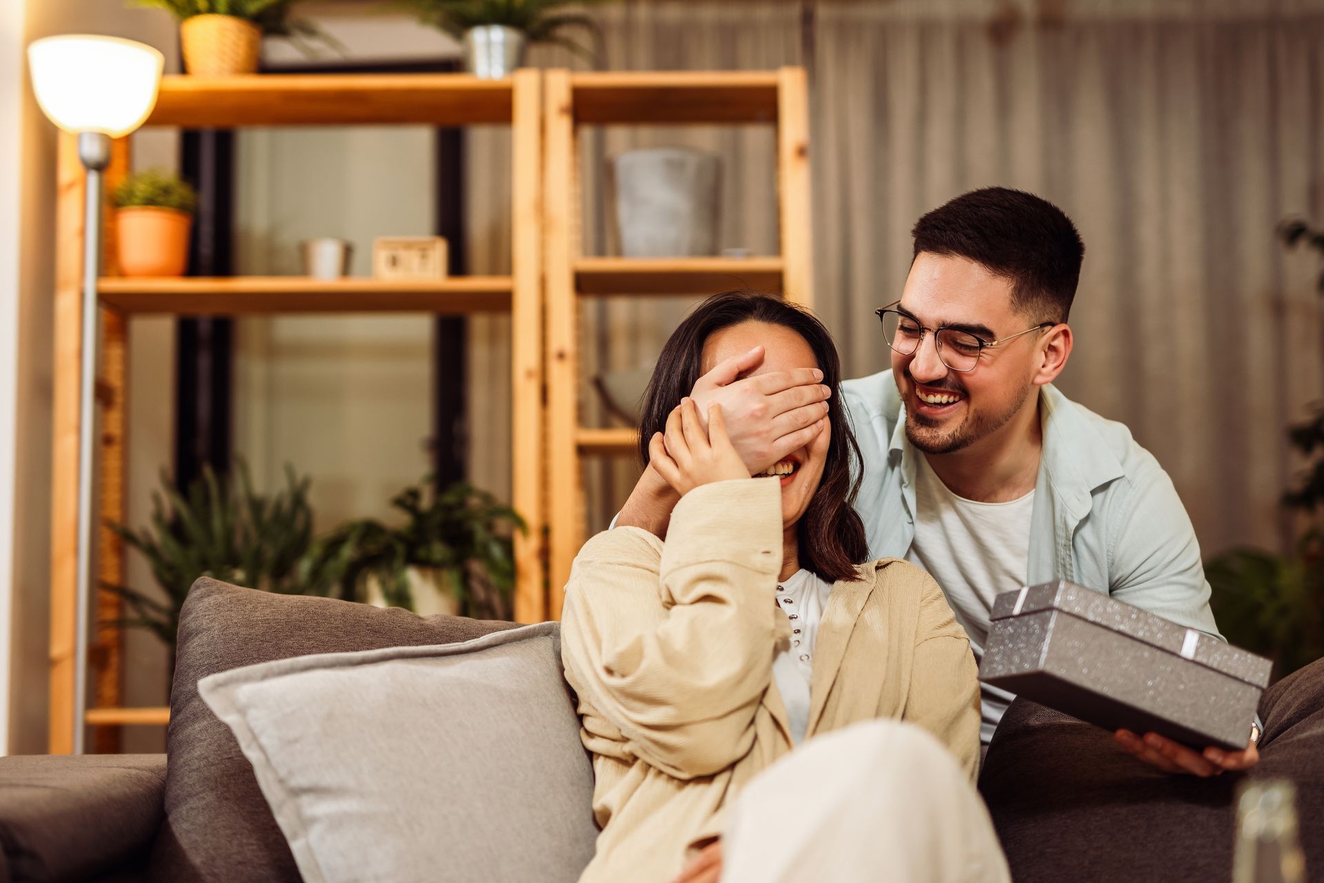 Man surprises woman with a gift, covering her eyes. They sit on a couch indoors.